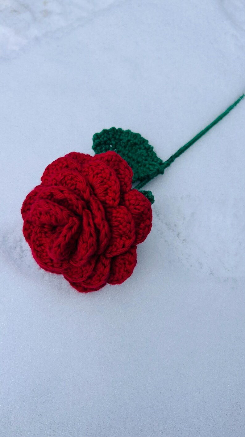 May include: A handmade, red crochet rose with a green leaf and stem. The flower is in full bloom, with detailed petals. The stem and leaf are a vibrant green. The rose is a craft item, possibly for decoration or as a gift.