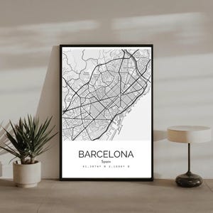 May include: Black and white map of Barcelona, Spain, in a black frame. The map shows the city's streets and landmarks. The word "Barcelona" is printed in a white rectangle at the bottom, along with the city's coordinates.
