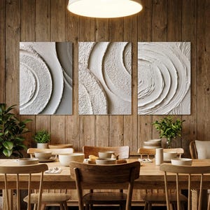 Puede incluir: Un tríptico de tres piezas de arte mural rectangular con pintura blanca gruesa y texturizada aplicada en patrones circulares y arremolinados. La obra muestra crestas elevadas y surcos profundos, creando un efecto táctil tridimensional.
