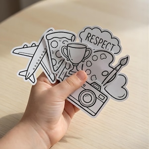 May include: A hand holding a collection of black and white stickers. The stickers include an aeroplane, a slice of pizza, a trophy, a cloud with the word "RESPECT", a paint palette with a brush, and a camera.