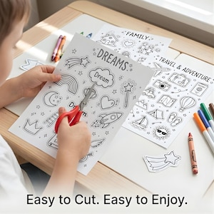 May include: A child uses red scissors to cut out a star from a black and white coloring sheet. The sheet is printed with the word "DREAMS" and various illustrations. Other sheets with illustrations and crayons are on the table. The text "Easy to Cut. Easy to Enjoy." is at the bottom.