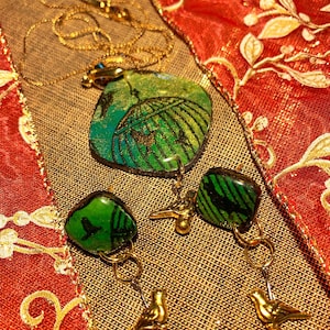 May include: A gold necklace with a pendant and matching earrings. The pendant and earrings feature green and blue stones with a leaf design. The earrings have gold bird charms.