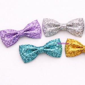 May include: Five glitter bow ties in various colors: purple, silver, black, teal, and gold. Each bow tie is made of a sparkly material and is tied in a classic bow shape. The bow ties are arranged on a white surface.
