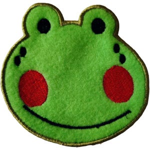 May include: A green frog-shaped patch with a gold embroidered border. The frog has black eyes, red cheeks, and a smiling black mouth. The patch is made of a soft, textured material.