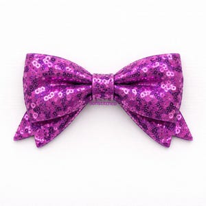 May include: A vibrant purple sequined bow, perfect for hair accessories or embellishments. The bow is centered on a white background, showcasing its sparkling texture and symmetrical design. The bow's color is a rich, eye-catching shade of purple.