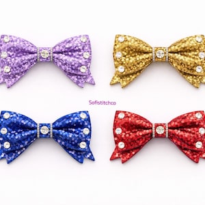 May include: Four sequined bow hair clips in purple, gold, blue, and red. Each bow features a rhinestone center and is set against a white background. The bows are likely for hair accessories.