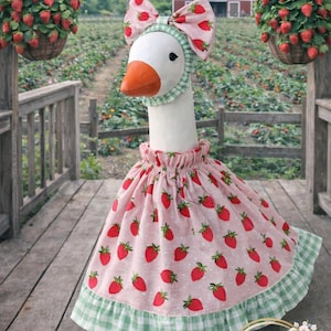 May include: A goose figurine wearing a pink dress with red strawberry patterns, a matching bow, and a green gingham trim. The goose is standing on a wooden deck with hanging baskets of strawberries in the background.