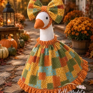 May include: A white goose figurine wearing a patchwork dress and a matching oversized bow headpiece. The fabric features a repeating pattern of orange, yellow, green, and teal squares with floral and polka dot prints. The dress has a ruffled hem.