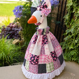 May include: A decorative goose statue with a white body, orange beak, and a patchwork dress in pink, white, and navy floral patterns. The goose wears a matching bonnet and bow. The dress has a white eyelet lace trim.
