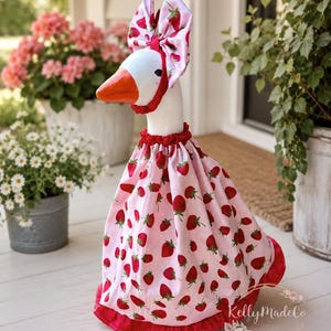 May include: A decorative goose figurine wearing a light pink dress featuring a red strawberry pattern and a matching red polka-dot ruffled hem. The goose also wears a coordinating strawberry-patterned headpiece with a red polka-dot trim.