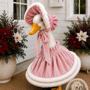 May include: A white goose statue wearing a pink velvet bonnet with white faux fur trim and a matching pink velvet skirt with a white faux fur hem. The goose has an orange beak and feet. A circular logo with the text Kelly Made & Company is visible.