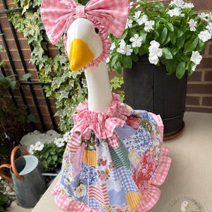 May include: A white plastic goose statue wearing a pink and white gingham bonnet with a large bow and a matching ruffled dress featuring a multi-colored patchwork floral pattern. The goose has a bright yellow beak and black eyes.