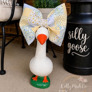 May include: A white goose statue with an orange beak and feet, perched on a green base. A large bow with yellow gingham and floral patterns adorns the goose's head. A black container with the words "silly goose" is in the background.