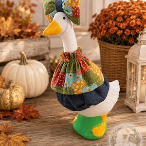 May include: A white ceramic goose figurine wearing a patchwork fabric outfit featuring floral patterns in green, red, and yellow, paired with a matching bow on its head and a dark blue denim-style skirt. The goose has a bright yellow beak and feet.