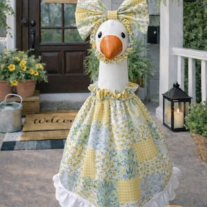 May include: A decorative goose statue wearing a yellow and white floral patterned dress and matching bow. The goose has a white body, orange beak, and is standing on a green base. The dress has a white eyelet lace trim.