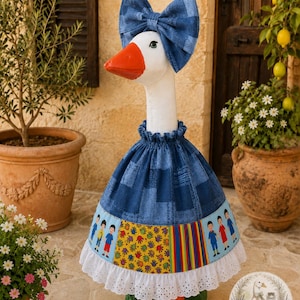 May include: A decorative goose figurine wearing a blue patchwork fabric outfit with a matching oversized bow on its head. The dress features a skirt with panels of children illustrations, colorful handprints, and vertical stripes, finished with a white eyelet lace trim.