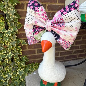 May include: A white goose statue with an orange beak and feet, and a large patchwork bow. The bow features floral, polka dot, and plaid patterns in pink, white, and navy blue. The goose stands on a green base.