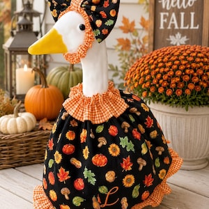 May include: A decorative goose figurine wearing a black fabric outfit featuring a pattern of pumpkins, sunflowers, mushrooms, and leaves. The outfit includes an orange and white gingham ruffled collar and hem, with a matching bonnet and an orange letter L.