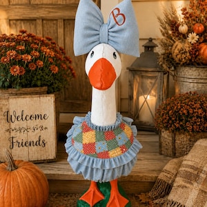 May include: A white goose figurine with an orange beak and feet, wearing a light blue denim bow with a brown letter B embroidered on it. The goose is dressed in a ruffled light blue denim collar and a multi-colored patchwork bib with floral patterns.