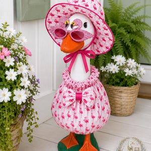 May include: A white goose statue wearing a pink flamingo-patterned fabric outfit consisting of a ruffled dress with a bow and a matching sun hat. The goose wears pink glittery flamingo-shaped sunglasses and has an orange beak and orange webbed feet.