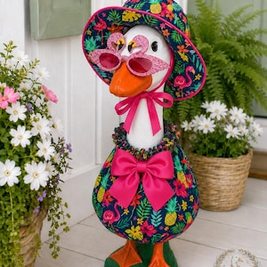 May include: A white goose statue wearing a dark blue bucket hat and matching dress with a tropical print of pink flamingos, yellow pineapples, and green leaves. The goose wears pink flamingo-shaped sunglasses and a large bright pink satin bow.