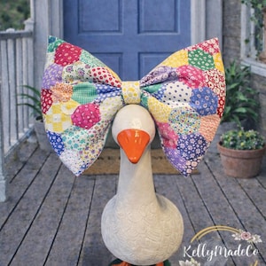 Może przedstawiać: Kapryśna statua gęsi z dużą, kolorową kokardą patchworkową. Gęś jest biała z pomarańczowym dziobem i stopami, stojąca na zielonej podstawie. Kokarda ma różne wzory i kolory tkanin. Tło obejmuje niebieskie drzwi i drewnianą werandę.
