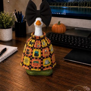 May include: A white goose figurine wearing a large black fabric bow on its head and a gathered skirt featuring a colorful Halloween-themed grid pattern with illustrations of black cats, cauldrons, ghosts, pumpkins, spiderwebs, and tombstones.