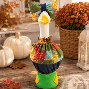 May include: A white goose figurine wearing a patchwork fabric outfit featuring sunflowers and a denim-like base. The goose has a bright yellow beak and feet, and is adorned with a matching oversized patchwork bow on its head and a ruffled collar.
