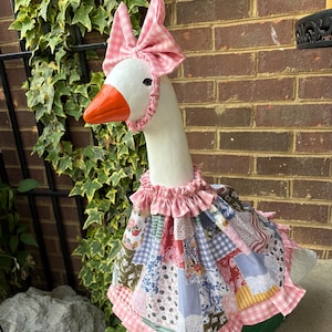 May include: A white goose statue with an orange beak, wearing a pink and white gingham bonnet with a matching ruffled collar and a patchwork dress featuring various floral, plaid, and solid fabric patterns in shades of pink, blue, and green.