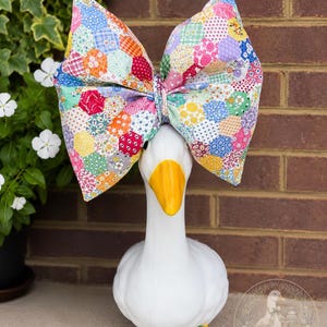 May include: A white goose figurine with a yellow beak wearing a large, oversized fabric bow. The bow features a colorful patchwork pattern made of various small hexagonal fabric pieces in shades of red, blue, yellow, pink, and green floral prints.