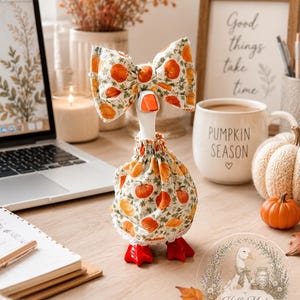 May include: A white goose figurine wearing a fabric dress and a matching oversized bow, both featuring a pattern of orange pumpkins and green foliage on a cream background. The goose has a bright orange beak and orange webbed feet.
