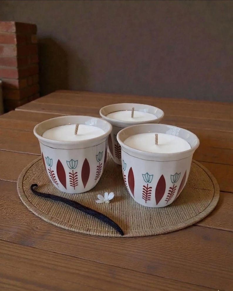 May include: Three white ceramic cups filled with white wax and a central wick. The exterior of each cup features a repeating pattern of dark red vertical leaf shapes and light blue tulip-like flowers. A single vanilla bean lies nearby.