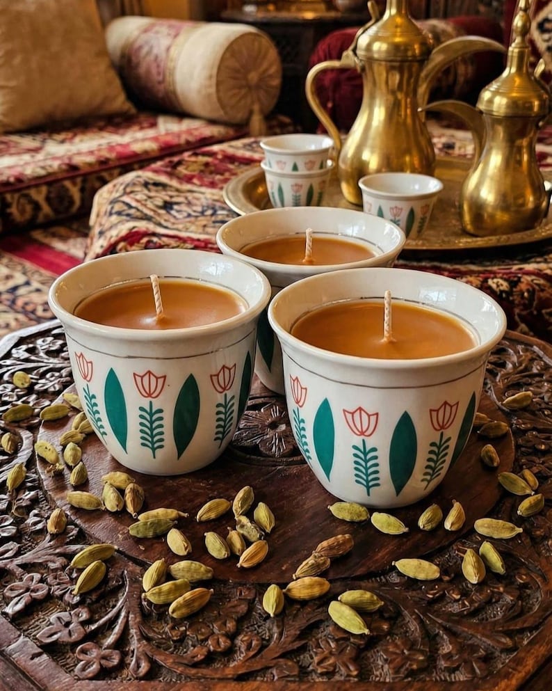 May include: Three white ceramic cups filled with orange wax and a central wick. Each cup features a repeating pattern of green leaves and orange tulip-like flowers. The cups are arranged on a dark carved wooden surface scattered with green cardamom pods.