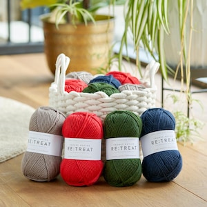 Peut inclure: Des pelotes de laine à tricoter épaisses et colorées dans un panier blanc tressé et sur une surface en bois. Les couleurs de la laine comprennent le gris, le rouge, le vert et le bleu marine. Chaque pelote porte une étiquette blanche avec le texte "RE:TREAT".