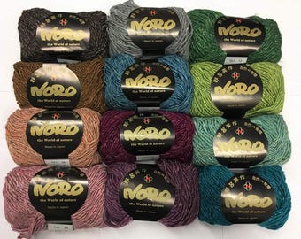 May include: A collection of Noro yarn skeins in various colors, including green, blue, brown, and purple. Each skein has a label with the brand name and the text "The World of Nature". The yarn is made in Japan.