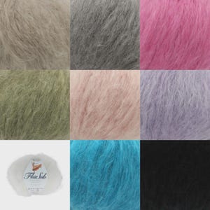 Op de afbeelding: Een collectie van negen bollen pluizig, getextureerd garen in diverse kleuren, waaronder beige, grijs, roze, olijfgroen, lichtroze, lavendel, wit, turquoise en zwart. Het witte garen heeft een label met de tekst Flair Solo Chunky.