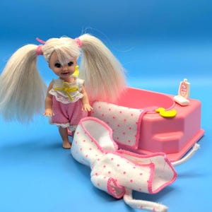 May include: A small doll with blonde hair in two pigtails, wearing a pink romper with a white lace collar and yellow ribbon. Beside the doll is a pink plastic bathtub, a white towel with pink dots, a yellow rubber duck, and a white soap bar.