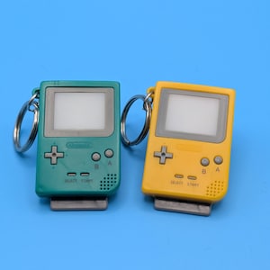 May include: Two miniature handheld game console keychains, one teal and one yellow, with silver key rings. Each has a screen, directional pad, buttons, and the text "SELECT START". The background is a solid blue.