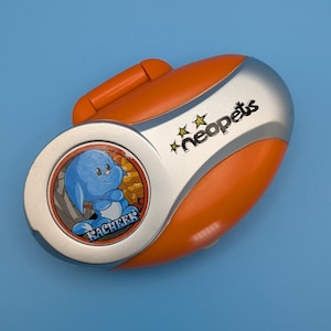 May include: An orange and silver Neopets handheld device featuring a cartoon Kacheek character. The device has a handle and the word "Kacheek" is printed on the round face. The Neopets logo is printed on the device.