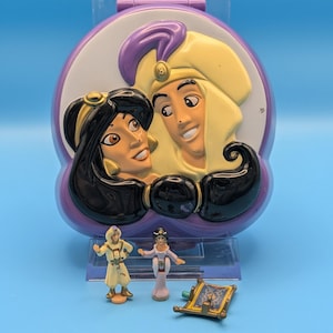 Op de afbeelding: Een vintage Aladdin-themaspeelgoedset. De set bevat een paarse en witte compact met Jasmine en Aladdin, samen met miniatuurbeeldjes van de personages en een vliegend tapijt. De achtergrond is effen blauw.