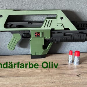 Könnte beinhalten: Eine zweifarbige, olivgrüne und schwarze Requisitenwaffe mit einer Digitalanzeige, die "34" anzeigt. Zwei silberne Patronen mit roten Deckeln liegen neben der Waffe. Der Text "Sekundärfarbe Oliv" steht unten.