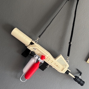 Op de afbeelding: Een crèmekleurig speelgoedpistool met een zwarte riem, rode en witte tanks en zwarte accenten. Het speelgoedpistool is aan een grijze muur bevestigd. Het pistool heeft een cilindrische loop en een rechthoekig lichaam met details.