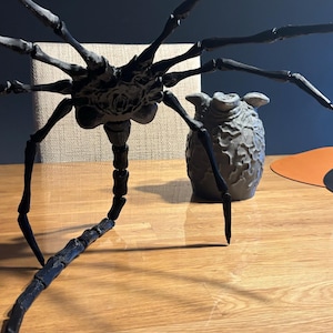 Pode incluir: Uma escultura de aranha preta articulada com múltiplas pernas e um corpo detalhado está em uma superfície de madeira. Um vaso cinza texturizado está ao lado da aranha. A cena é montada em uma mesa de madeira.