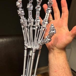 Könnte beinhalten: Eine detaillierte, silberfarbene Roboterhand mit beweglichen Fingern. Die mechanische Hand ist eine Nachbildung, die komplizierte Gelenke und metallische Komponenten zeigt. Die Hand wird zum Größenvergleich gegen eine menschliche Hand gehalten.