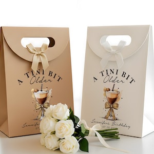 Könnte beinhalten: Zwei Geschenktüten, eine beige und eine cremefarbene, mit Griff und Schleife. Jede Tüte trägt den Text "A Tini Bit Older" und eine Cocktail-Illustration. Ein Strauß weißer Rosen mit weißem Band liegt vor den Tüten.