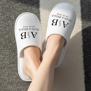 Op de afbeelding: Witte badstof slippers met de tekst "AB" en de namen "ALAN & BELLA" samen met de datum "05.22.2026" in zwart gedrukt. De slippers liggen op een zwart-wit gestreept tapijt.