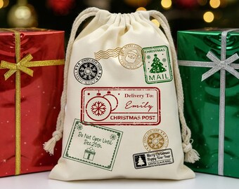 May include: A cream-colored Christmas gift bag with a drawstring closure, adorned with festive stamps. The stamps include a Santa Claus image, a Christmas tree, and the text "Delivery To: Emily, Christmas Post." Beside the bag are wrapped gifts.