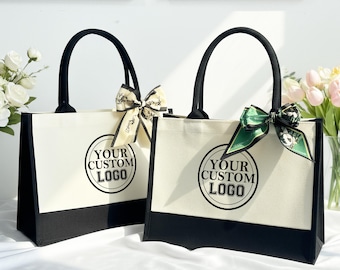 May include: Two canvas tote bags with black handles and bottoms. Each bag has a white body with a black border and the text "YOUR CUSTOM LOGO" in a black circle. One bag has a cream-colored bow, and the other has a green bow.