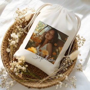 May include: A cream drawstring bag featuring a photograph of a person amidst flowers. The bag is placed in a woven basket, decorated with dried flowers. The name "Sophia" is written in script on the bag.
