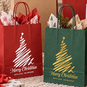 Puede incluir: Una bolsa de regalo de papel rojo con un árbol de Navidad garabateado en blanco y el texto Merry Christmas WITH LOVE, THE BROWNS, y una bolsa de regalo de papel verde con un árbol de Navidad garabateado en dorado y el texto Merry Christmas WITH LOVE, THE BROWNS.